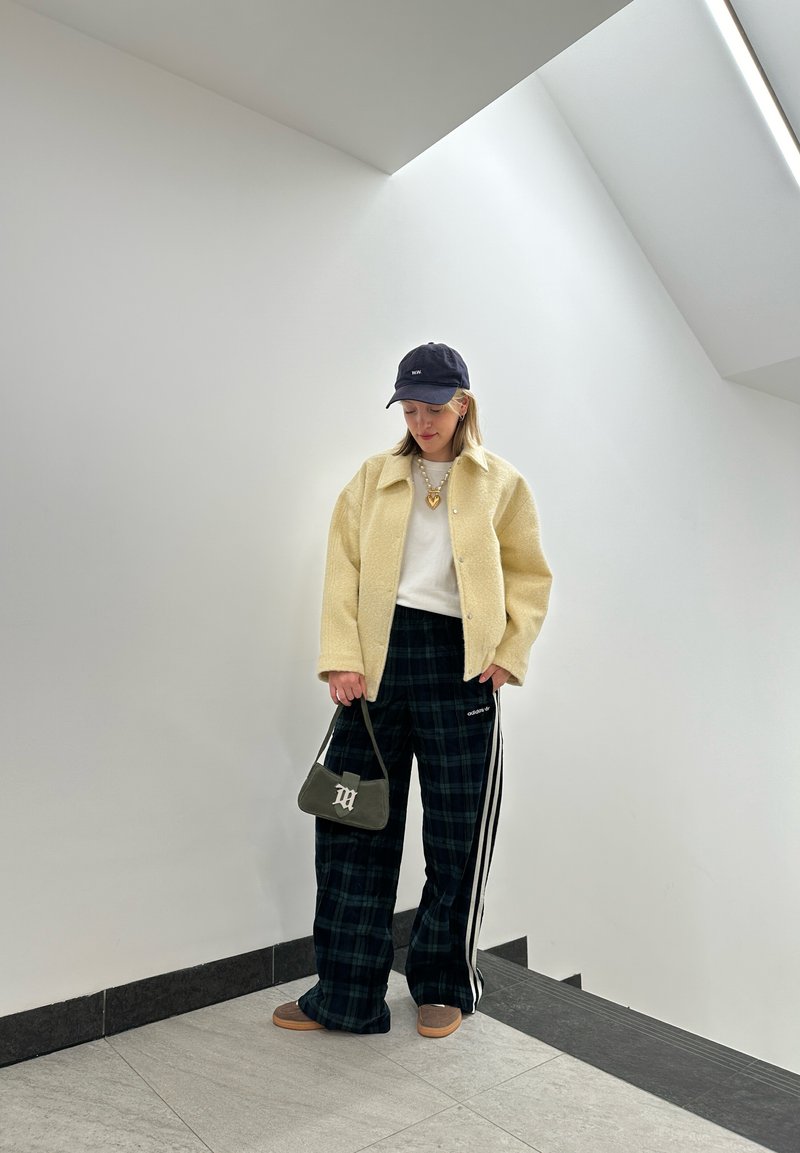 Κίτρινο υφασμάτινο μπουφάν, λευκή μπλούζα, καρό παντελόνι σε φαρδιά γραμμή με ρίγες, μαύρο καπέλο, πράσινη τσάντα χειρός και καφέ παπούτσια σε φωτεινό, μινιμαλιστικό περιβάλλον.