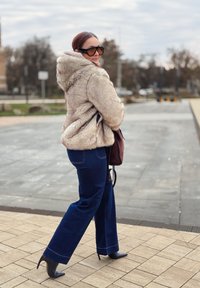 Jacke aus Kunstpelz in hellem Beige, oversized mit Kapuze; marineblaue ausgestellte Jeans mit Seitentaschen; schwarze Stilettoabsätze; braune Handtasche.