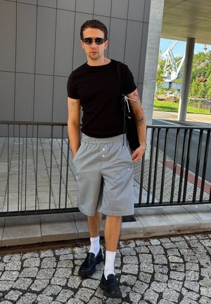 Camiseta negra ajustada, pantalones cortos grises a cuadros con cintura elástica, calcetines blancos, mocasines negros y un bolso negro de hombro.