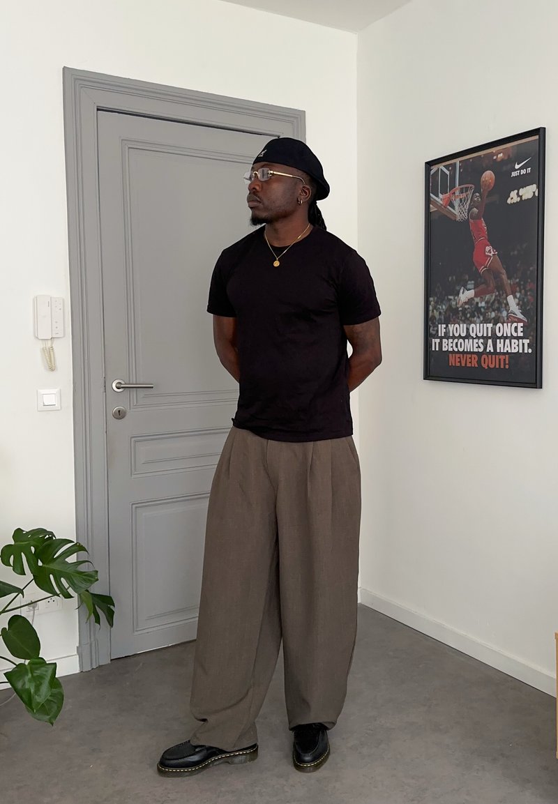 Mann trägt schwarzes Hemd, graue weite Hose und rückwärts getragenen Cap, steht mit den Händen hinter dem Rücken nahe einer grauen Tür und einem motivierenden Basketballposter.