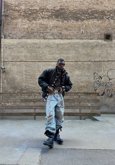 Hombre con gafas de sol, chaqueta negra, vaqueros rotos y botas negras se apoya en un banco de madera en un entorno urbano con grafitis en la pared.