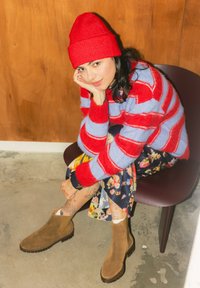 Jeune femme aux cheveux noirs portant un bonnet rouge, un pull rayé rouge et bleu, une jupe à fleurs et des bottines en daim marron, assise sur une chaise à l'intérieur.