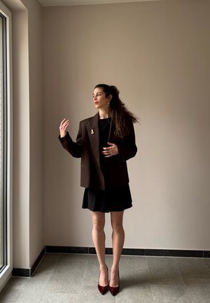 Mujer con vestido negro y blazer marrón oversize está junto a una ventana, mirando hacia un lado con una mano levantada y la otra en la cintura.