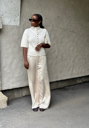 Mujer con top crema abotonado, pantalones anchos de satén, zapatos oscuros, gafas de sol y sujetando un pequeño bolso de mano, de pie frente a una pared texturizada.