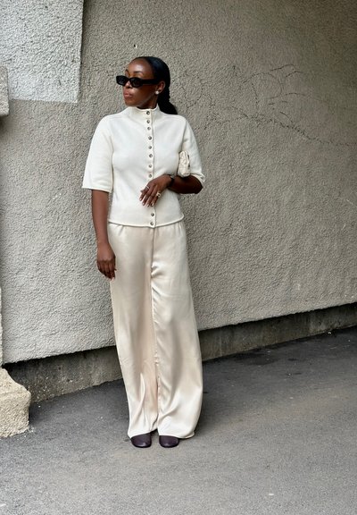Mujer con top crema abotonado, pantalones anchos de satén, zapatos oscuros, gafas de sol y sujetando un pequeño bolso de mano, de pie frente a una pared texturizada.
