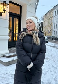 Svart polstringet kåpe med hette, hvit strikket lue og en beige rutete skjerf. Hun står på snødekte trinn ved siden av en bygning.