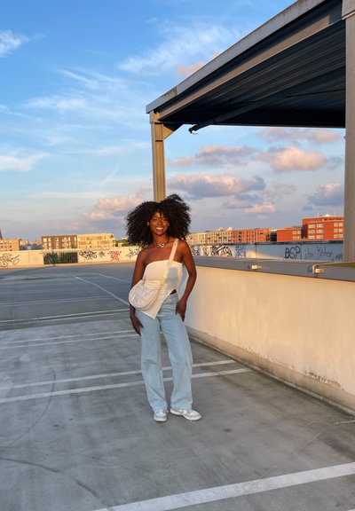 Top blanco de canalé con un solo hombro y jeans de pierna ancha de color azul claro. Se lleva un bolso pequeño de un color claro, cruzado sobre el cuerpo. Fondo de aparcamiento en una azotea urbana.