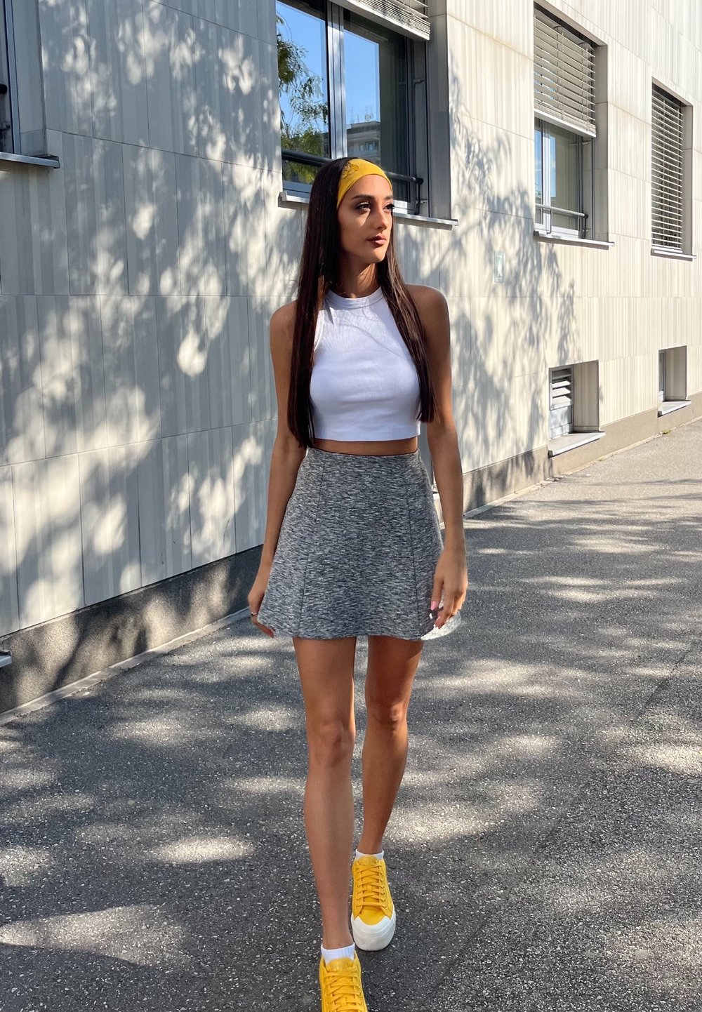 Γκρι φαρδύ σορτς, λευκό κομμένο μπλουζάκι και φωτεινές κίτρινες αθλητικές παπούτσια. Μακριά μαλλιά χτενισμένα με μια κίτρινη κορδέλα. Αστική φόντο με παράθυρα.