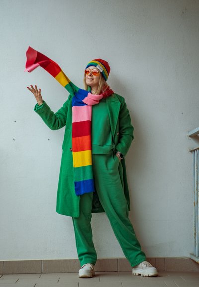 Persona con abrigo y pantalones verdes que lleva una bufanda y un gorro a rayas de colores del arcoíris, gafas de sol naranjas y zapatos blancos, de pie contra una pared lisa.