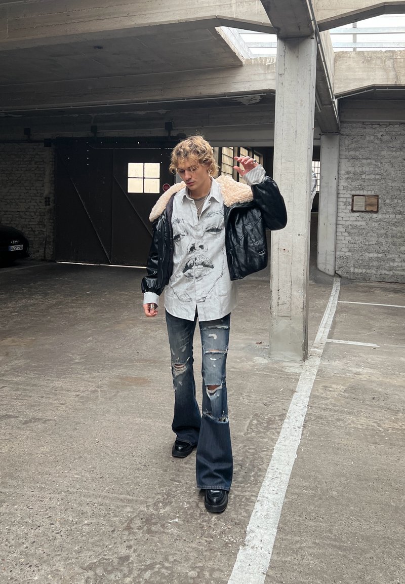 Black leather jacket with shearling collar, oversized white shirt with graphic print, ripped dark jeans, black shoes, posing in a concrete space.