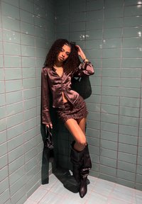 Braune Satinbluse mit geraffter Front, kombiniert mit einem Pailletten-Minirock. Das Model trägt schwarze kniehohe Stiefel und hält eine schwarze Tasche.