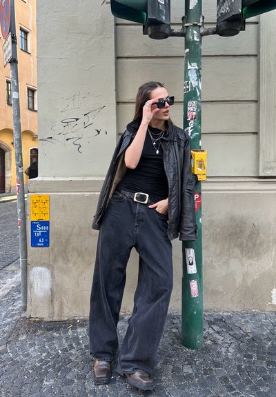 Chaqueta de cuero negra sobre una camiseta negra, combinada con vaqueros negros de pierna amplia y botas marrones. Los accesorios incluyen collares de plata en capas y gafas de sol.