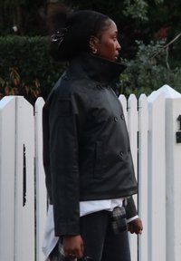 Svart skinnjakke med dobbelspent lukking og høy krage, med sidelommer og en teksturert finish, båret over en hvit skjorte og rutete bukse.