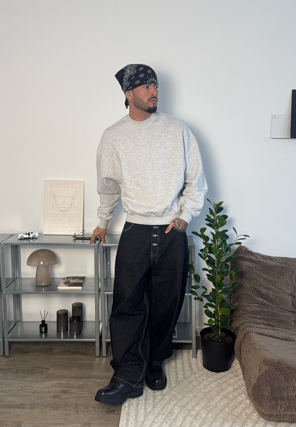 Mann trägt ein graues Sweatshirt, ein schwarzes Bandana und weite schwarze Jeans und steht neben Metallschränken und einer Topfpflanze in einem Wohnzimmer.