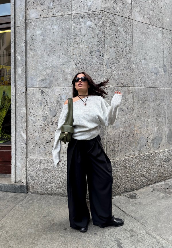 Une femme portant des lunettes de soleil, un pull gris et un pantalon noir à jambes larges pose près d’un mur en pierre, avec un sac à bandoulière vert olive.