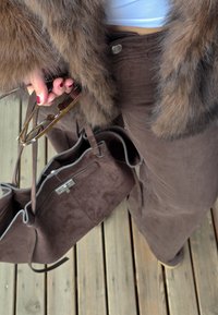 Eine Hand, die eine Schildpattbrille und eine braune Wildlederhandtasche hält, trägt braune Cordhosen und einen Pelzmantel und steht auf einem Holzboden.