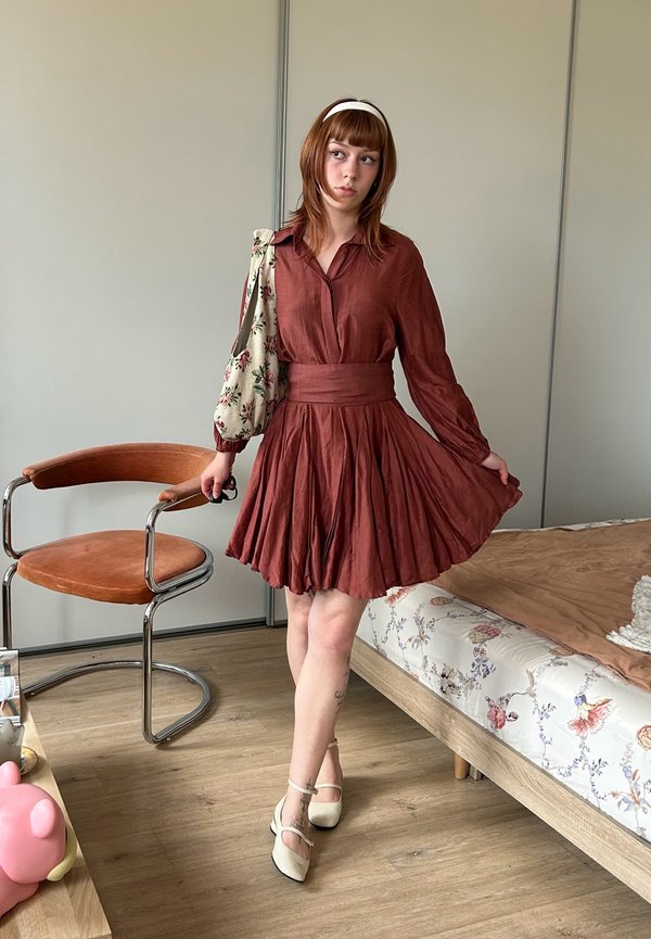 Joven mujer con un vestido marrón plisado y zapatos blancos está de pie junto a una cama y una silla de madera en un dormitorio minimalista.