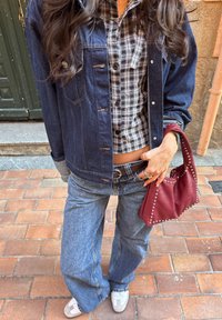 Casaco de ganga, camisa xadrez preto e branco, jeans azuis largos, bolsa bordô com tachas, sapatilhas brancas, colocado num fundo de rua de tijolos.