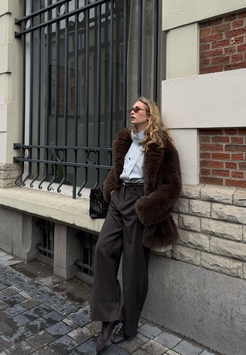 Mujer con abrigo de piel marrón, pantalones negros de pierna ancha y gafas de sol apoyada contra una pared de piedra con una ventana con rejas de hierro en la acera de la ciudad.