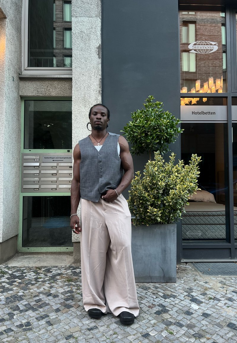 Γκρι γιλέκο πάνω από ένα λευκό πουκάμισο, κρεμ παντελόνι με φαρδύ πόδι και μαύρες παπούτσια. Άνδρας στέκεται δίπλα σε έναν μικρό θάμνο κοντά σε μια είσοδο κτιρίου.