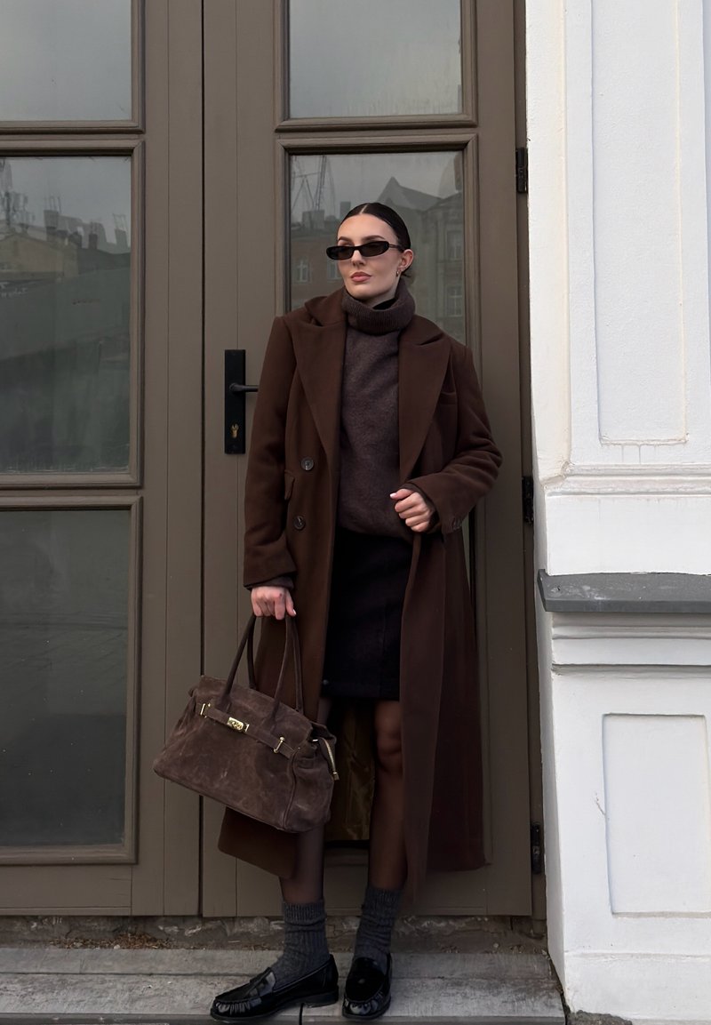 Donna con cappotto lungo marrone, gonna nera, dolcevita scuro, occhiali da sole, che tiene una borsa marrone, in piedi davanti a una porta doppia chiusa in un contesto urbano.