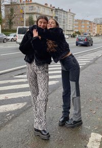 Fekete műszőrme dzseki, zebra mintájú, bő nadrág, és sötét farmer világos foltokkal. Vastag fekete platform cipők. Városi utcai környezet.