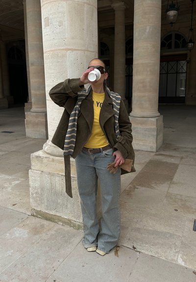 Persona con gafas de sol, camisa amarilla, chaqueta oliva, bufanda a rayas y vaqueros bebe de una taza mientras está de pie junto a columnas de piedra en un patio.