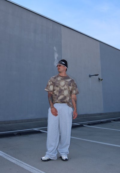 Camiseta de manga corta, tie-dye en tonos marrones y beige, combinada con pantalones de chándal gris claro. El atuendo se complementa con zapatillas blancas y un gorro negro.