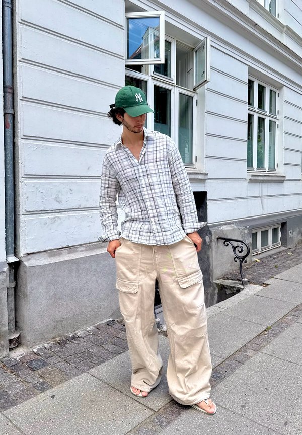Jeune homme en pantalon beige large, chemise à carreaux boutonnée, casquette verte et tongs, debout sur le trottoir urbain près d'un bâtiment blanc avec une fenêtre ouverte.