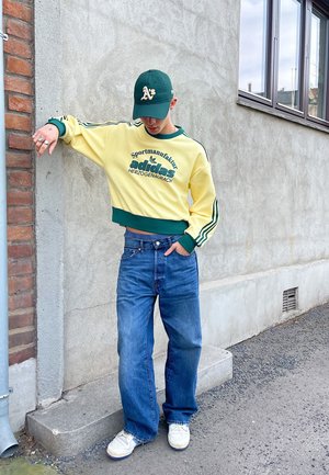 Sudadera amarilla corta con acentos y logotipo verdes, jeans anchos azules, zapatillas blancas y una gorra verde con texto. Fondo urbano sencillo.