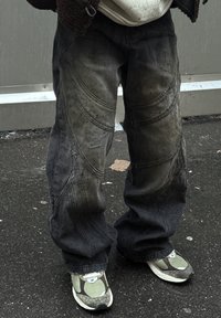 Juodos, platėjančios džinsinės kelnės su matomais purvo ir drėgnų dėmių pėdsakais, su išlenktomis siūlėmis ir laisvu kirpimu. Dėvimos su pilkai žaliais sportbačiais.