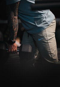 Vyras su tatuiruotu ranka kelia juodą šešiakampį hantelį, vilkėdamas mėlyną marškinėlį, smėlinės spalvos "Nike" kelnes ir juodą laikrodį sporto salėje.