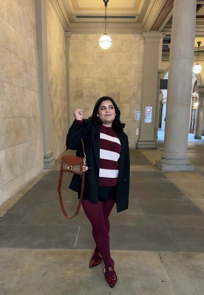 Blazer oscuro sobre un suéter a rayas burdeos y blanco, leggings granate y zapatos de punta con tachuelas. Bolso marrón con un acento dorado.
