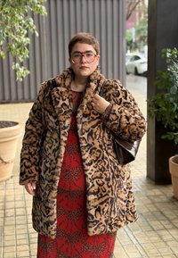 Personne aux cheveux courts portant de grandes lunettes, un manteau en fausse fourrure à motif léopard, une robe rouge à motifs, et tenant un sac à main marron foncé en plein air.