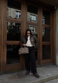 Giacca di pelle marrone sopra un maglione crema, abbinata a pantaloni neri e scarpe platform scure. Una borsa grigia con dettagli arricciati completa il look.