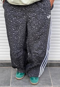 Calças de treino pretas com padrão de estampa de leopardo, apresentando riscas laterais brancas e um logótipo da Adidas em branco no lado esquerdo.