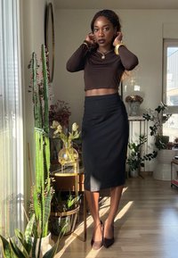 Femme ajustant ses cheveux, portant un crop top marron à manches longues, une jupe midi noire et des talons, debout dans une pièce éclairée par le soleil avec des plantes et un miroir rond.