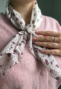 Personne portant un pull rose et une écharpe crème nouée avec des motifs floraux bordeaux, montrant des ongles manucurés et une bague délicate.
