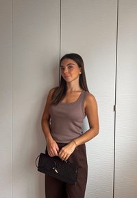 Bruine geribde tanktop gecombineerd met donkerbruine broeken; een zwart gewatteerd clutch in de hand. Lichte textuurachtergrond in zachte tinten.