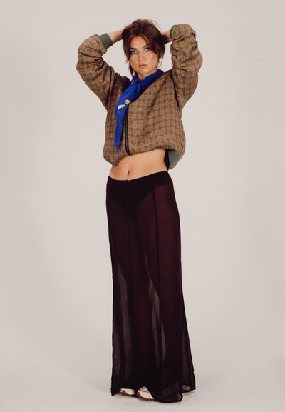 Chaqueta bomber con estampado en tonos tierra, combinada con una falda maxi negra transparente. Una bufanda azul añade un toque de color. La modelo posa con las manos levantadas.