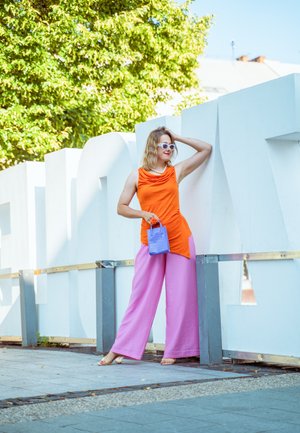 Top sin mangas naranja con escote drapeado, pantalones de pierna ancha rosa y un pequeño bolso azul. Los accesorios incluyen gafas de sol blancas.