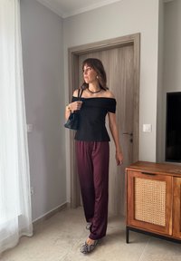 Sieviete stāv telpās pie koka durvīm, valkājot melnu krekliņu bez pleciem, bordo satīna bikses, čūskas raksta kurpes un turot melnu somiņu.