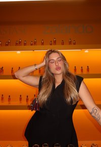 Svart ärmlös klänning bärs av en person framför en livlig orange bakgrund med glasflaskor av hårprodukter på hyllor.