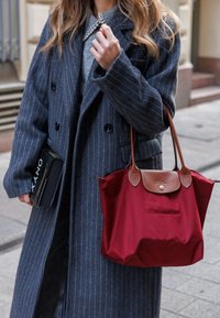 Punainen nylon-kassi, jossa on ruskeita nahkaosuuksia, pidetään tummanharmaan raidallisen takin vieressä. Mustaa lehteä puristetaan toisessa kädessä.