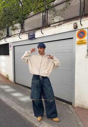 Sudadera de cremallera de color beige claro con grandes bolsillos, combinada con jeans amplios de mezclilla azul oscuro desgastados que presentan dobladillos deshilachados. Unos zapatos beige completan el look.