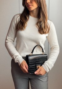 Žena v bílém svetru a šedých kalhotách, držící černou kabelku s krokodýlím vzorem a zlatým zapínáním před texturovanou zdí.