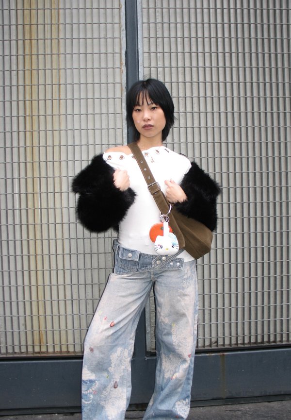 Person with short black hair wears white top with black furry sleeves, bag with plush keychain, and baggy patterned jeans standing against metal grid.