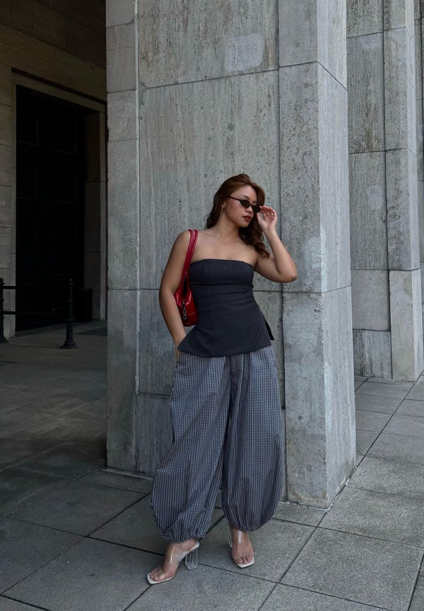 Žena v čiernom top bez ramienok a voľných kockovaných nohaviciach si upravuje slnečné okuliare, stojí pri vysokých kamenných stĺpoch vonku.