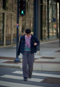 Avslappnad klädsel med en mörk jacka över en genomskinlig topp med blommig broderi, vida byxor med stort mönster och svarta skor, som går på övergångsställe.