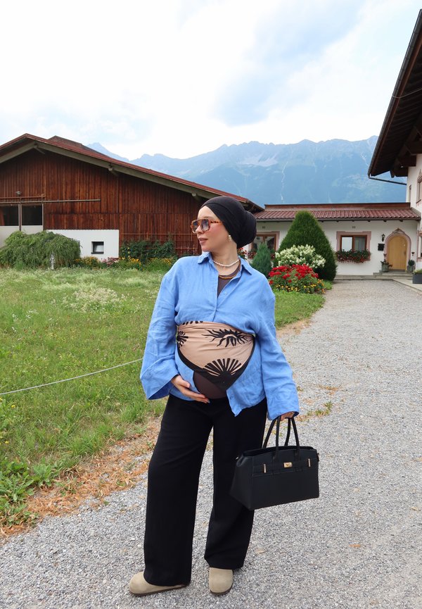 Femme enceinte portant un foulard et des lunettes de soleil, chemise bleue, pantalon noir, tenant un sac à main noir, debout sur un chemin de gravier près de fleurs et d'une maison en bois.